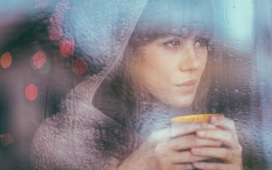 Kvinna med kopp i handen bakom ett fönster med regndroppar på.