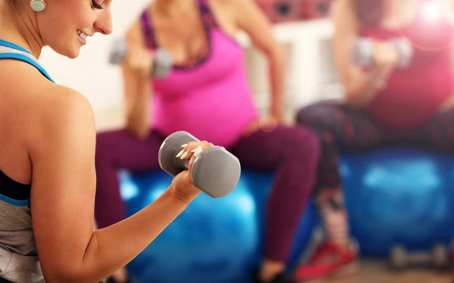 Gravid kvinna sitter på pilatesboll och gör bicepsövning med hantel.